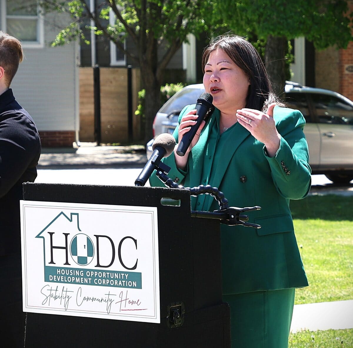 An Asian woman wearing green speaks at a podium labeled "HODC" outdoors on a sunny day, with white woman wearing black using sign language standing next to her. Green lawn and trees in the background.