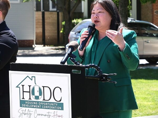 An Asian woman wearing green speaks at a podium labeled "HODC" outdoors on a sunny day, with white woman wearing black using sign language standing next to her. Green lawn and trees in the background.