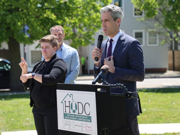 Man in suit speaks at a podium labeled "HODC" outdoors. A sign language interpreter in black translates. The mood is formal and focused.