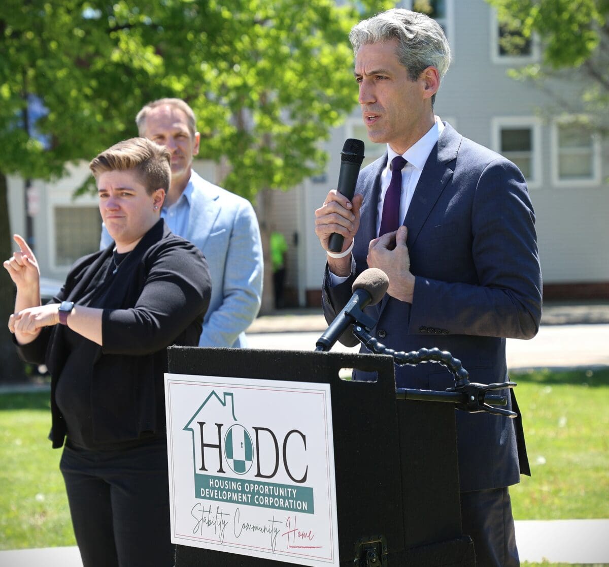 Man in suit speaks at a podium labeled "HODC" outdoors. A sign language interpreter in black translates. The mood is formal and focused.