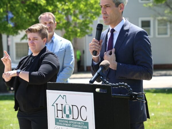 Man in suit speaks at a podium labeled "HODC" outdoors. A sign language interpreter in black translates. The mood is formal and focused.