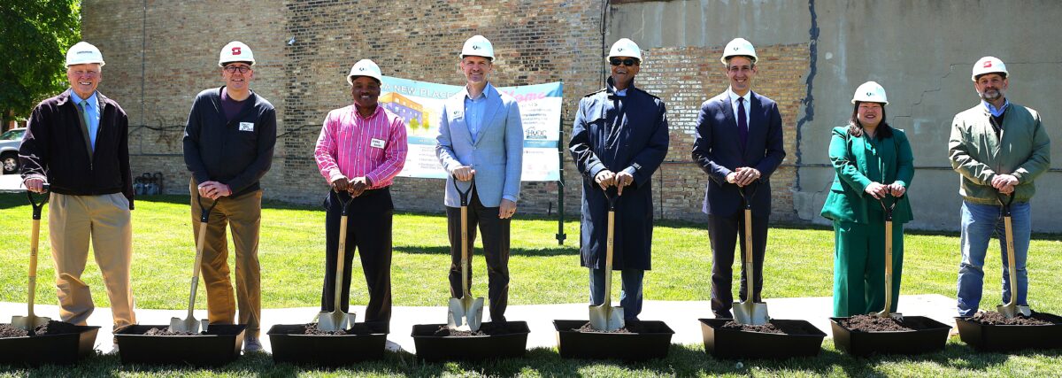 8 professionally dressed men and women wearing white hard hats pose with shovels for a groundbreaking ceremony. They smile, conveying a sense of accomplishment and teamwork.
