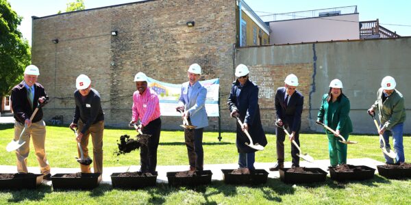 A group of eight people in hard hats participate in a groundbreaking ceremony. They are smiling and using shovels to turn soil, with a brick building backdrop.