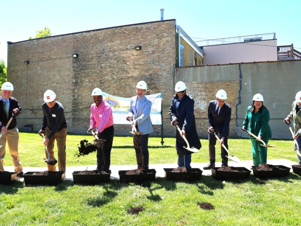 A group of eight people in hard hats participate in a groundbreaking ceremony. They are smiling and using shovels to turn soil, with a brick building backdrop.