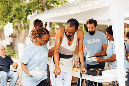 A woman carries a container of food while helping a man using two crutches. People are serving food under a white tent in the background.