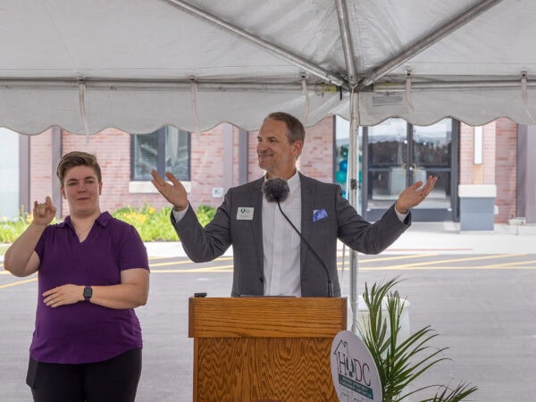 A man wearing a white shirt and a grey suit jacket gestures as he speaks from behind a podium. A woman with short hair and a purple shirt interprets in sign language next to him. They are standing under a white tent with a building in the background.