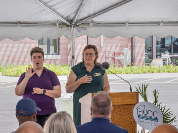 A woman wearing a blue sleeveless dress speaks from a podium. A woman wearing a purple shirt stands next to her interpreting in sign language. People are sitting in front of them listening.