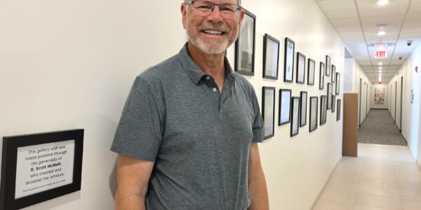 A white man wearing glasses and a grey short-sleeved shirt poses with a wall of framed pictures that is in the hallway of an apartment building.