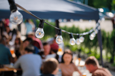 A line of outdoor party lights hang above a blurred image of people sitting under a party tent.