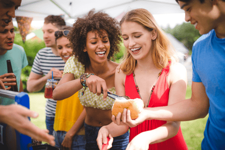 People at a BBQ under a tent. One woman is putting a hot dog into a bun held by a man.