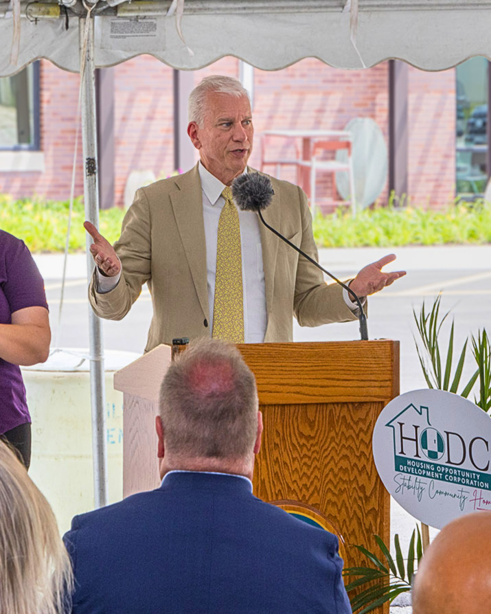 Scott Britton, wearing a tan suit and yellow tie, speaks from behind a podium as the audience listens.