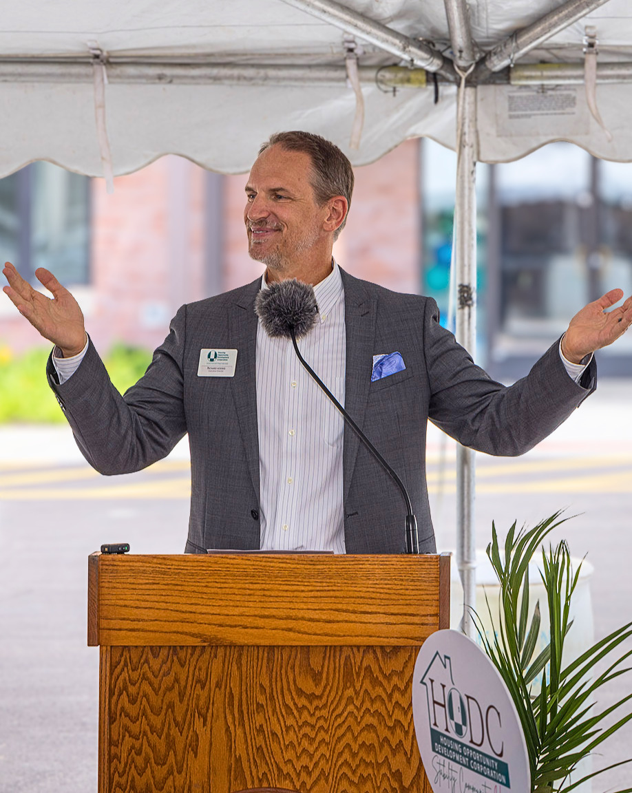 A man wearing a white button-down shirt and a grey suit jacket poses behind a podium with his arms raised in a welcoming manner.