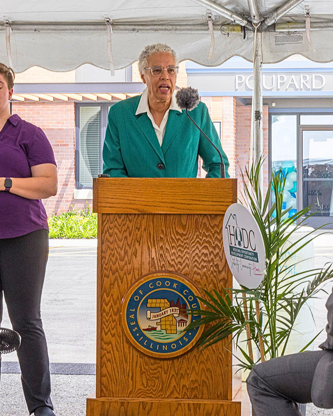 Toni Preckwinkle, wearing a green suit and white blouse, speaks from behind a podium with the Cook County seal.