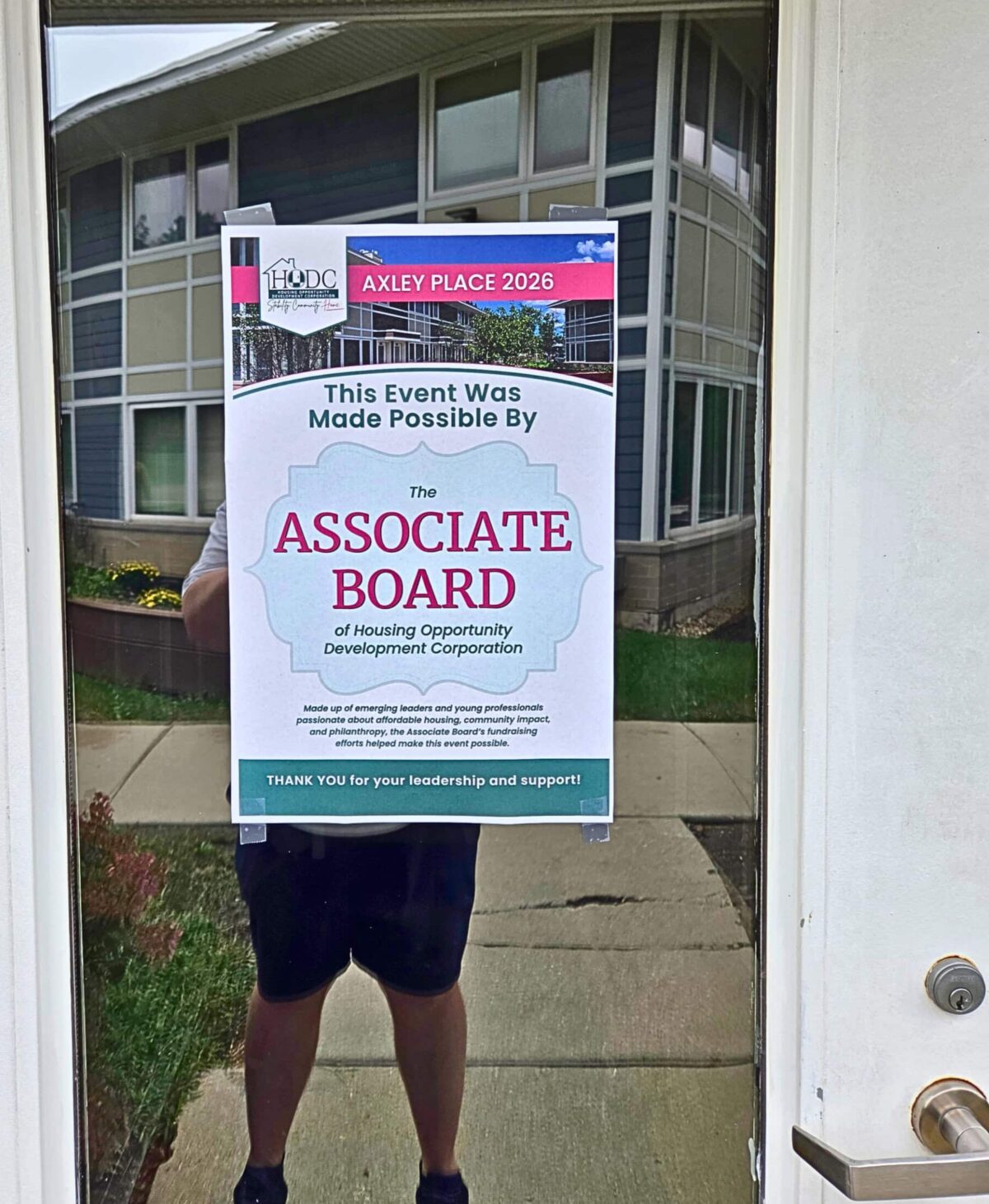 A poster saying that this event was made possible by HODC's Associate Board is taped to the glass of a door. An apartment building is reflected in the glass.