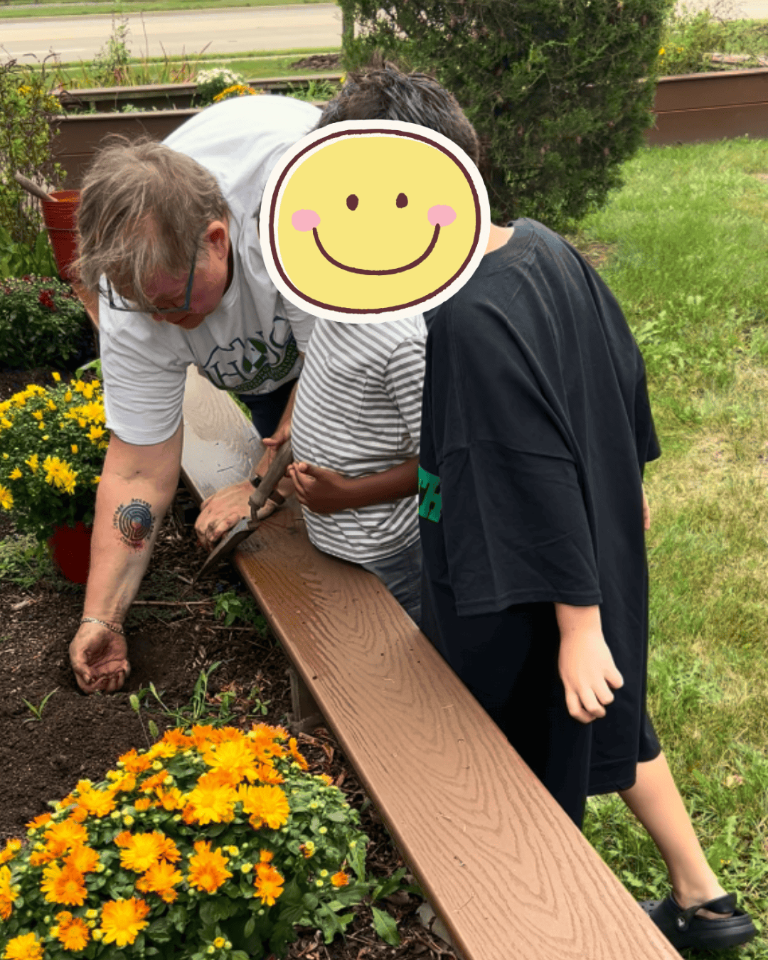 A woman bends as she digs a hold in a raised garden bed. Two children are standing next to her with one holding a small garden trowel.