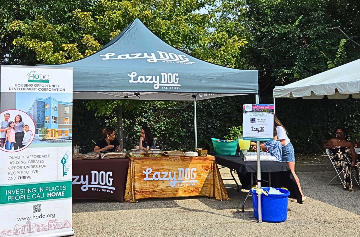 Lazy Dog Restaurant tent and tables with food. People are sitting behind the table. It is a sunny summer day.